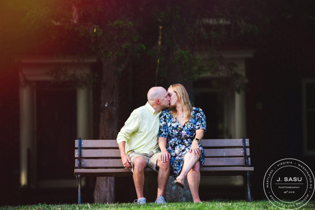 a couple cuddles and kisses while sitting on a bench in a park in Gaithesrburg, MD during their engagement session.